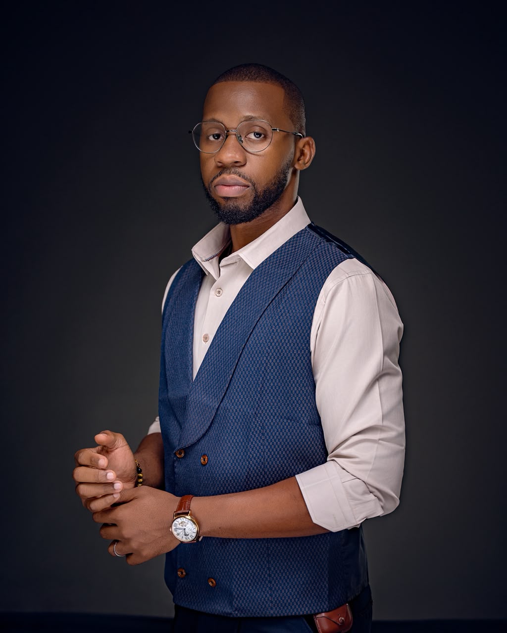 Zubby Gabriel standing against a dark grey studio wall in a navy waistcoat and cream shirt, hands clasped, wearing round glasses.